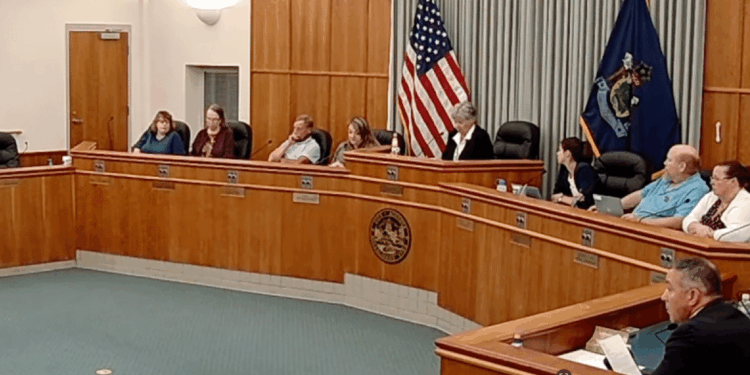 City council meeting in progress with members seated at a wooden table, American flag displayed, discussing local governance issues.