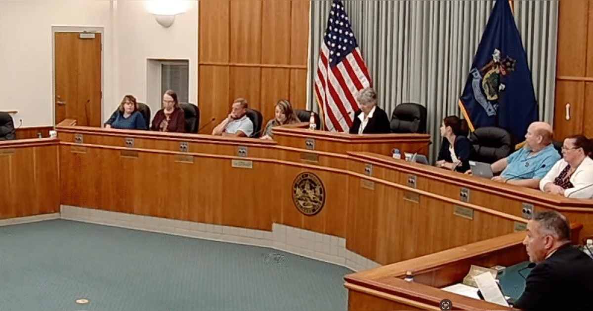 City council meeting in progress with members seated at a wooden table, American flag displayed, discussing local governance issues.