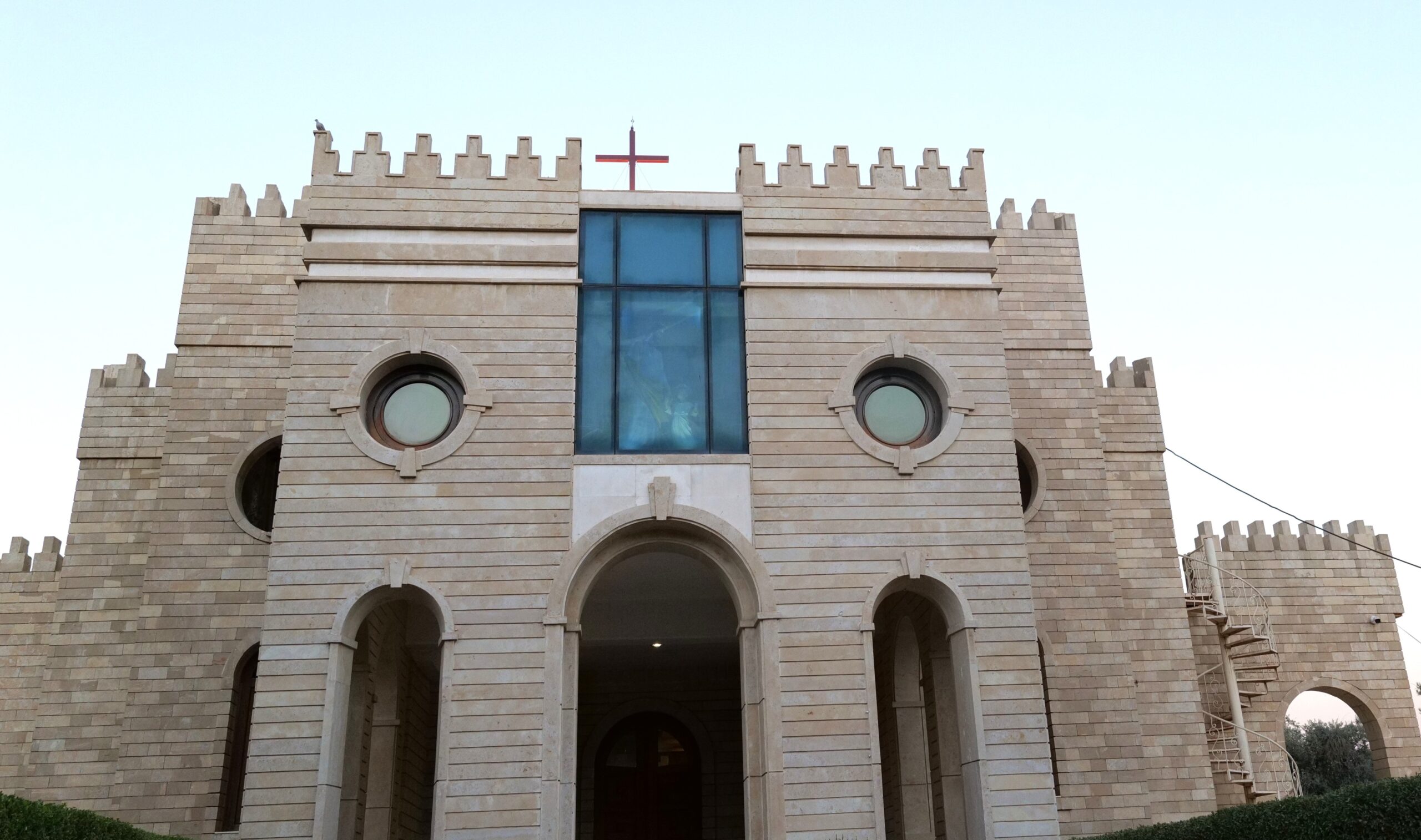 Historic stone building with medieval architecture and circular windows, featuring a prominent cross atop the structure against a clear sky.