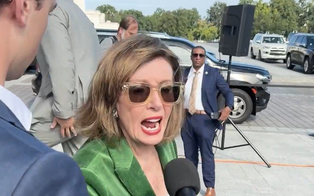 Nancy Pelosi speaking to the media while surrounded by security and vehicles, showcasing her engagement in a public event.