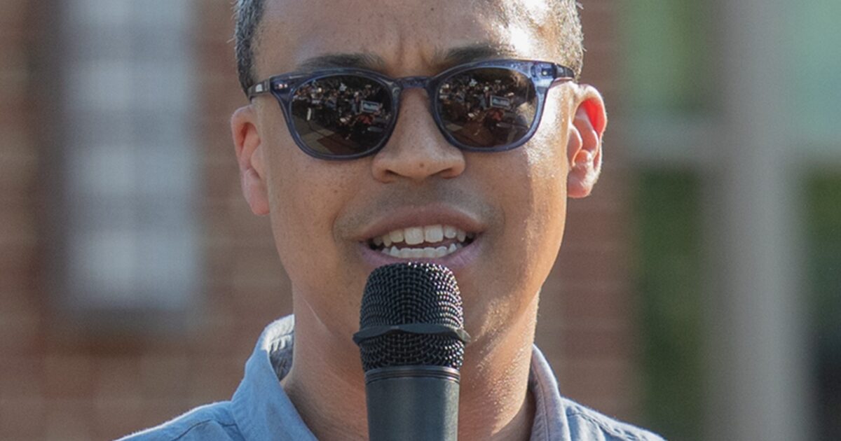 Man speaking into a microphone while wearing sunglasses, with a blurred crowd in the background during an outdoor event.
