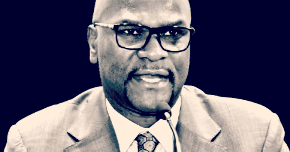 Business professional speaking at a conference, wearing glasses and a suit, engaged in a discussion with a microphone in front of him.