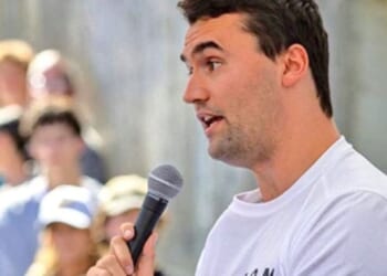 Young man speaking into a microphone at an outdoor event, engaging with an attentive crowd in the background.