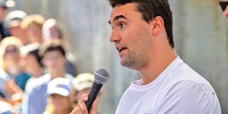Young man speaking into a microphone at an outdoor event, engaging with an attentive crowd in the background.