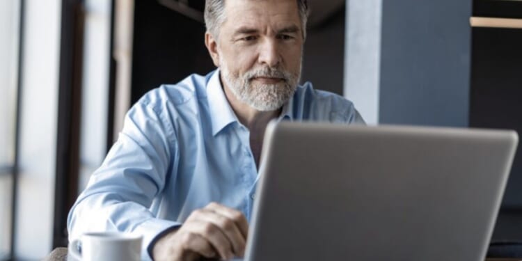 Middle-aged man with a beard working on a laptop in a modern office setting, focused on his task with a coffee cup nearby.