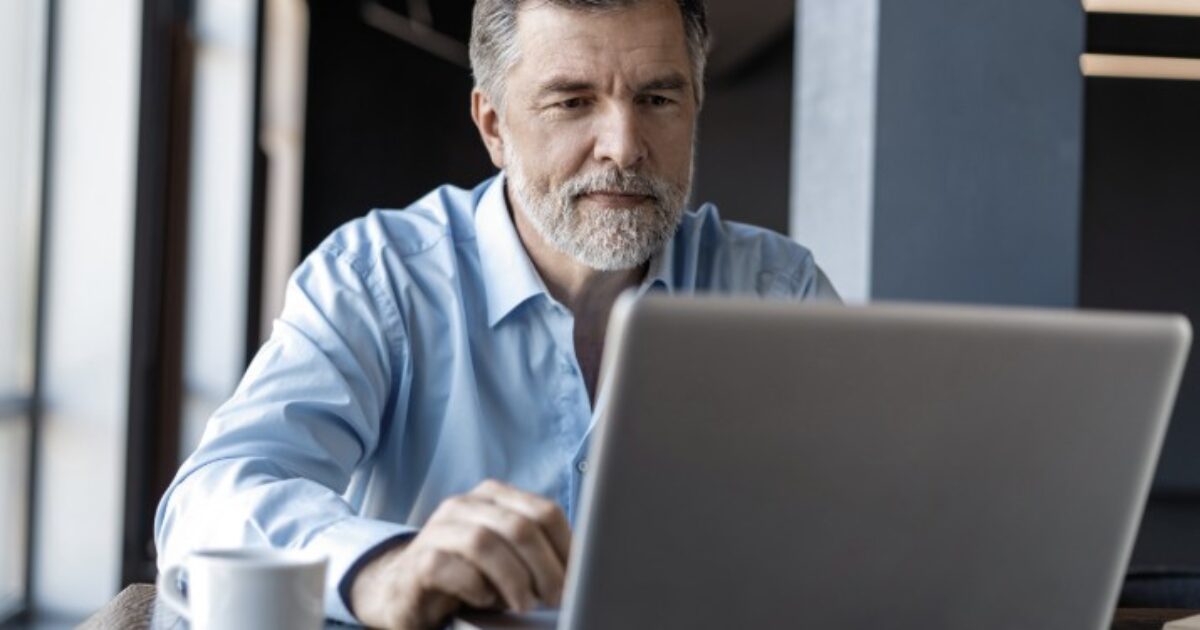 Middle-aged man with a beard working on a laptop in a modern office setting, focused on his task with a coffee cup nearby.
