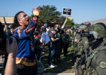 Protester passionately gestures during a demonstration against police presence, with law enforcement in tactical gear visible in the background.