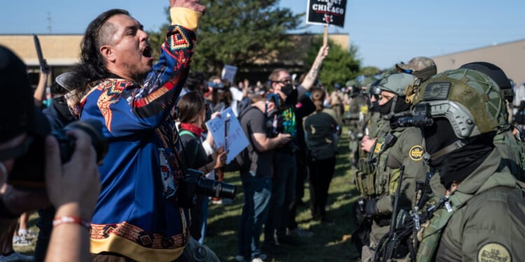 Protester passionately gestures during a demonstration against police presence, with law enforcement in tactical gear visible in the background.