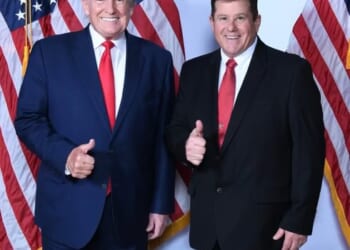 Donald Trump and a supporter pose together with thumbs up in front of American flags, highlighting a moment of camaraderie at a political event.