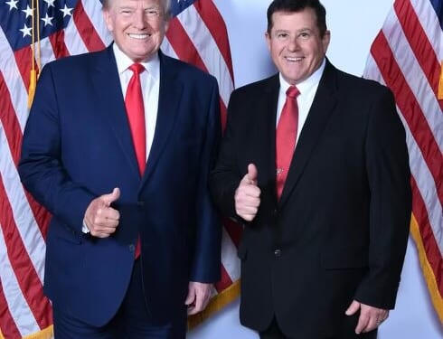 Donald Trump and a supporter pose together with thumbs up in front of American flags, highlighting a moment of camaraderie at a political event.