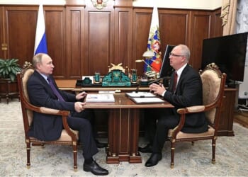 Vladimir Putin and a government official engage in a discussion at a formal meeting in an office setting, surrounded by Russian symbols and decor.