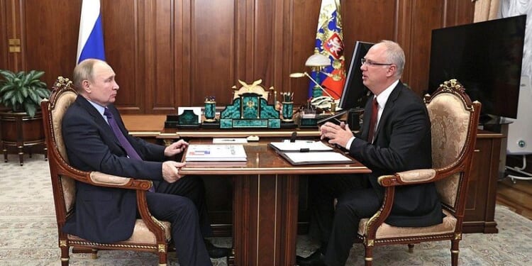 Vladimir Putin and a government official engage in a discussion at a formal meeting in an office setting, surrounded by Russian symbols and decor.