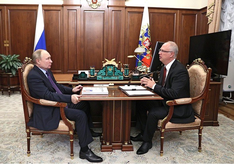 Vladimir Putin and a government official engage in a discussion at a formal meeting in an office setting, surrounded by Russian symbols and decor.