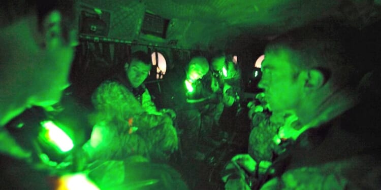Soldiers seated inside a military aircraft, illuminated by green lights, preparing for a night operation.