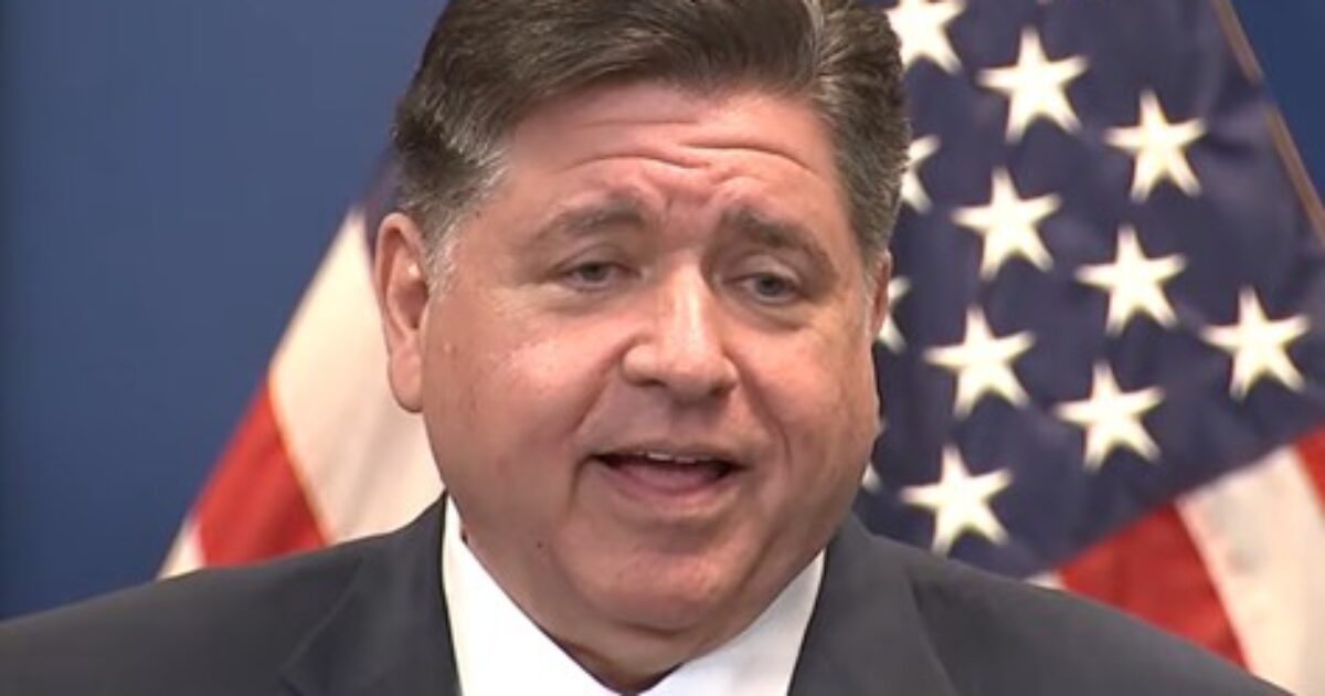 Illinois Governor JB Pritzker speaking at a press conference with American flags in the background.