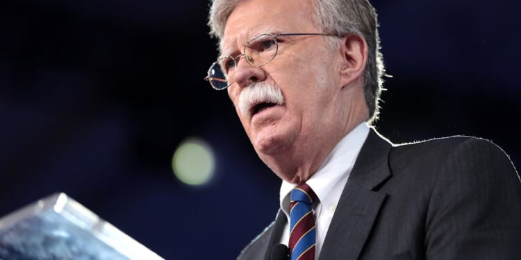 John Bolton speaking at a podium during a public event, emphasizing key political points with a serious expression.