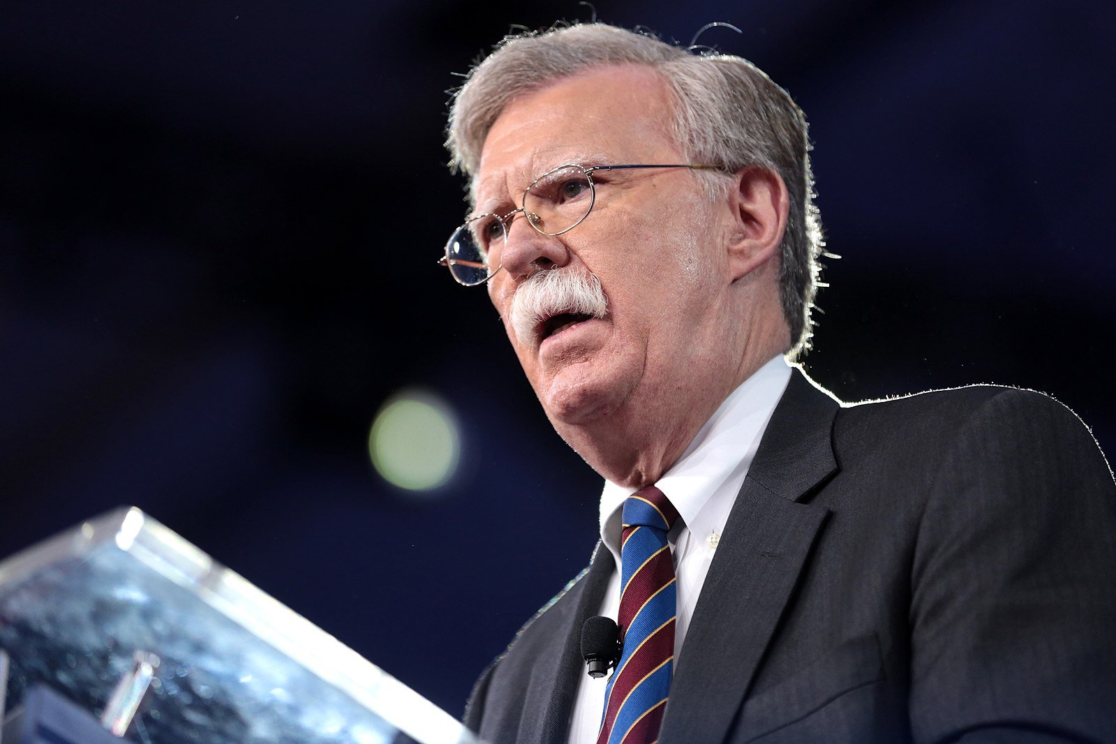 John Bolton speaking at a podium during a public event, emphasizing key political points with a serious expression.