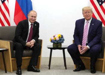 Russian President Vladimir Putin and former U.S. President Donald Trump meet for a discussion, with national flags in the background and a floral arrangement on the table.