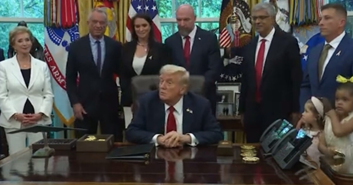 Donald Trump seated at a desk in the Oval Office surrounded by a diverse group of individuals during an official event.