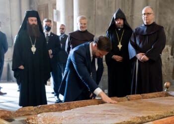 A group of clergy members observes a man in a suit touching a historic stone slab in a religious setting.