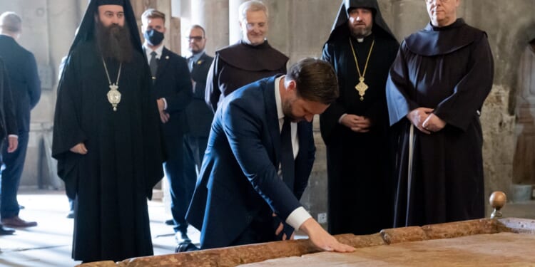 A group of clergy members observes a man in a suit touching a historic stone slab in a religious setting.