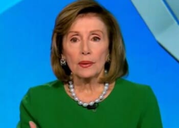 Female politician wearing a green dress and pearl necklace, speaking during a televised interview against a blue background.