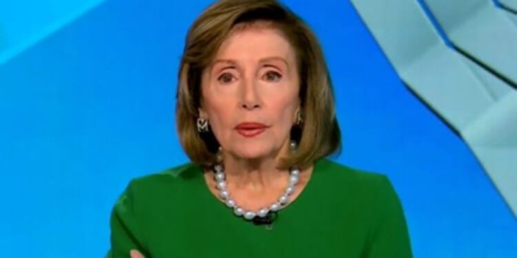 Female politician wearing a green dress and pearl necklace, speaking during a televised interview against a blue background.