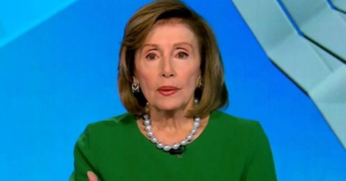 Female politician wearing a green dress and pearl necklace, speaking during a televised interview against a blue background.