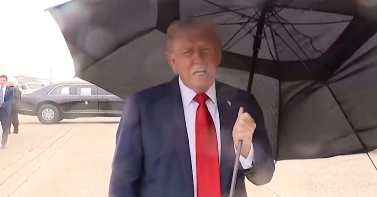 Donald Trump holding an umbrella while speaking to the press on a rainy day near a black presidential vehicle.