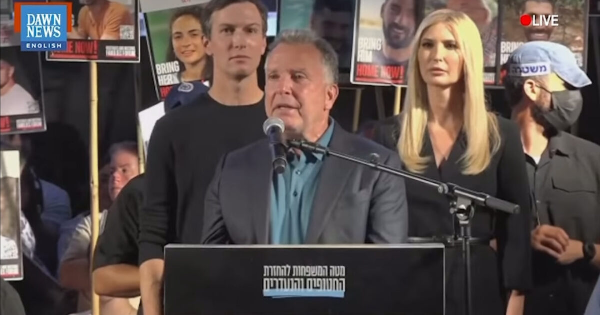 A speaker addresses a crowd during a live event, flanked by supporters holding signs advocating for the return of loved ones.