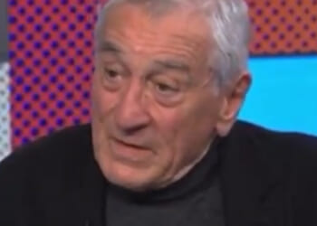 An older man with gray hair speaks thoughtfully during an interview, dressed in a black jacket against a colorful background.