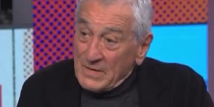 An older man with gray hair speaks thoughtfully during an interview, dressed in a black jacket against a colorful background.