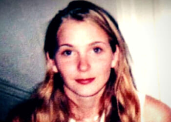 Young woman with long, wavy blonde hair and a neutral expression, posing in a softly lit indoor setting.