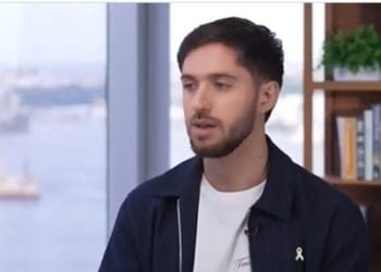 Young man with a beard speaking in an interview setting, with a cityscape visible in the background and a bookshelf beside him.