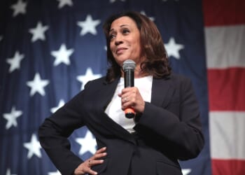 Kamala Harris speaking at a podium with a microphone, set against a backdrop of the American flag.