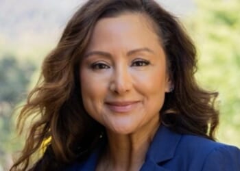Professional woman with long wavy hair smiling outdoors in a blue blazer against a blurred natural background.