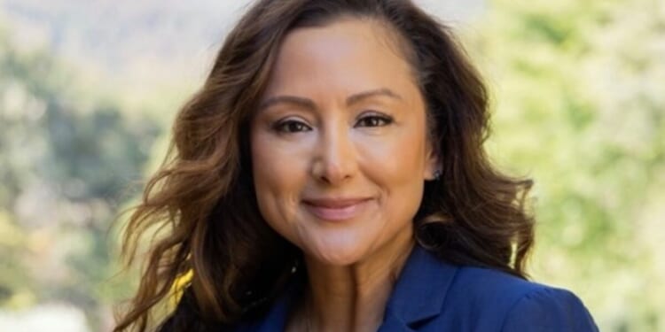 Professional woman with long wavy hair smiling outdoors in a blue blazer against a blurred natural background.