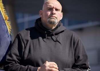 Man in a black hoodie stands confidently with hands clasped, framed by a flag, during an outdoor event in bright sunlight.