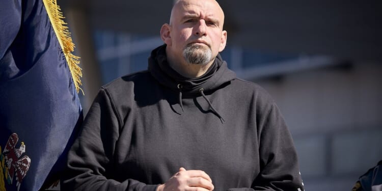 Man in a black hoodie stands confidently with hands clasped, framed by a flag, during an outdoor event in bright sunlight.