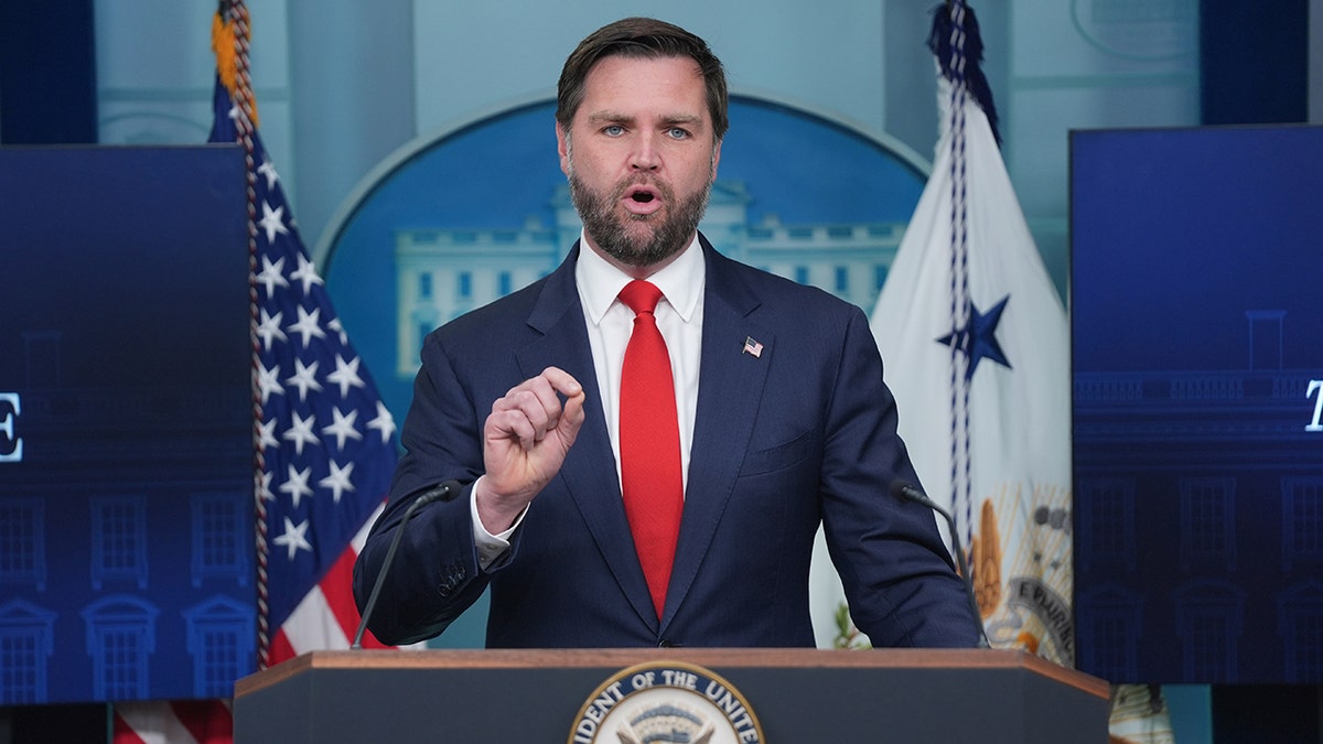 Vice President JD Vance wearing a dark suit and red tie while speaking at a podium in the White House
