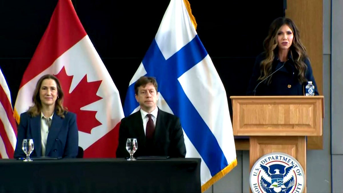 Kristi Noem speaking at a podium as Kirsten Hillman and Sakari Puisto sit off to the side