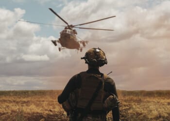 Soldier in tactical gear observing military helicopter landing in open field