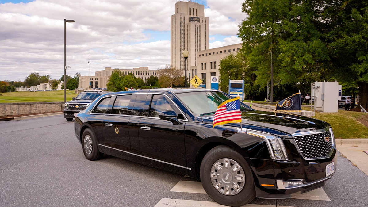 A motorcade vehicle carrying the president approaches a waiting military helicopter outside a medical facility.