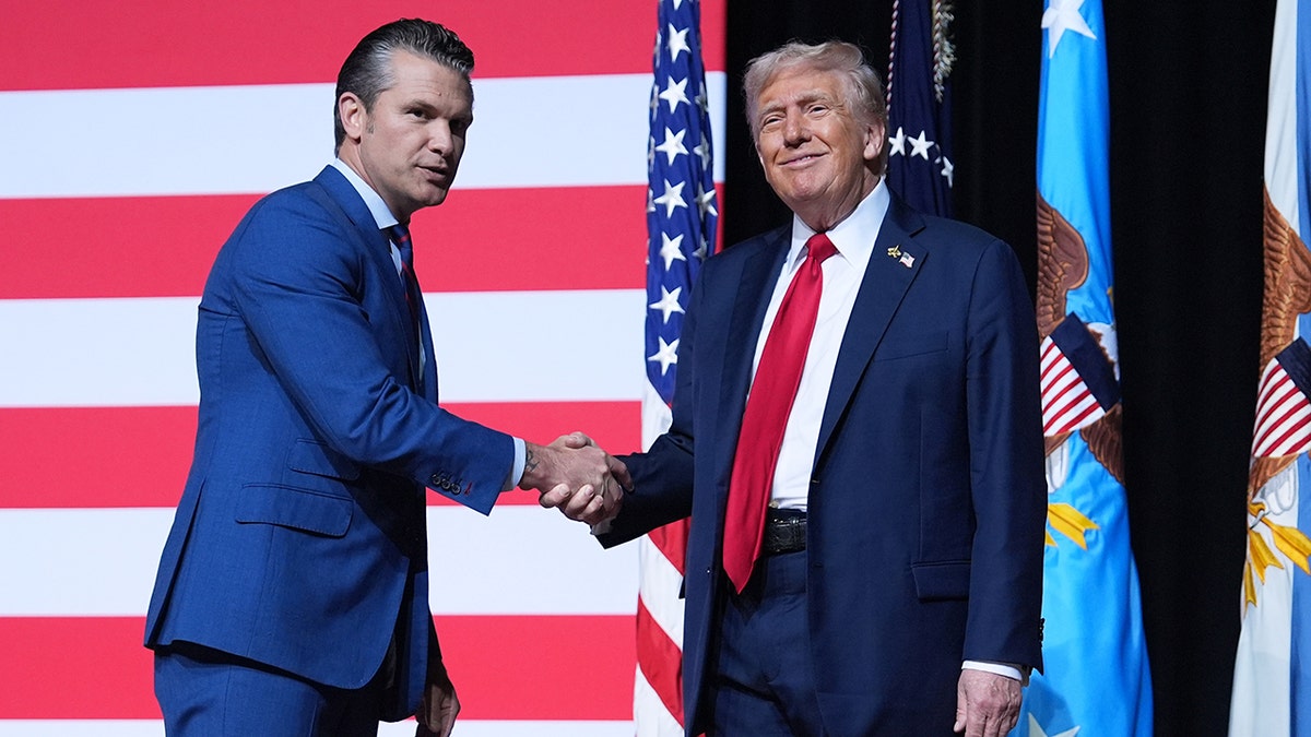 Pete Hegseth and Donald Trump, both wearing navy suits, shaking hands in front of American flag on stage