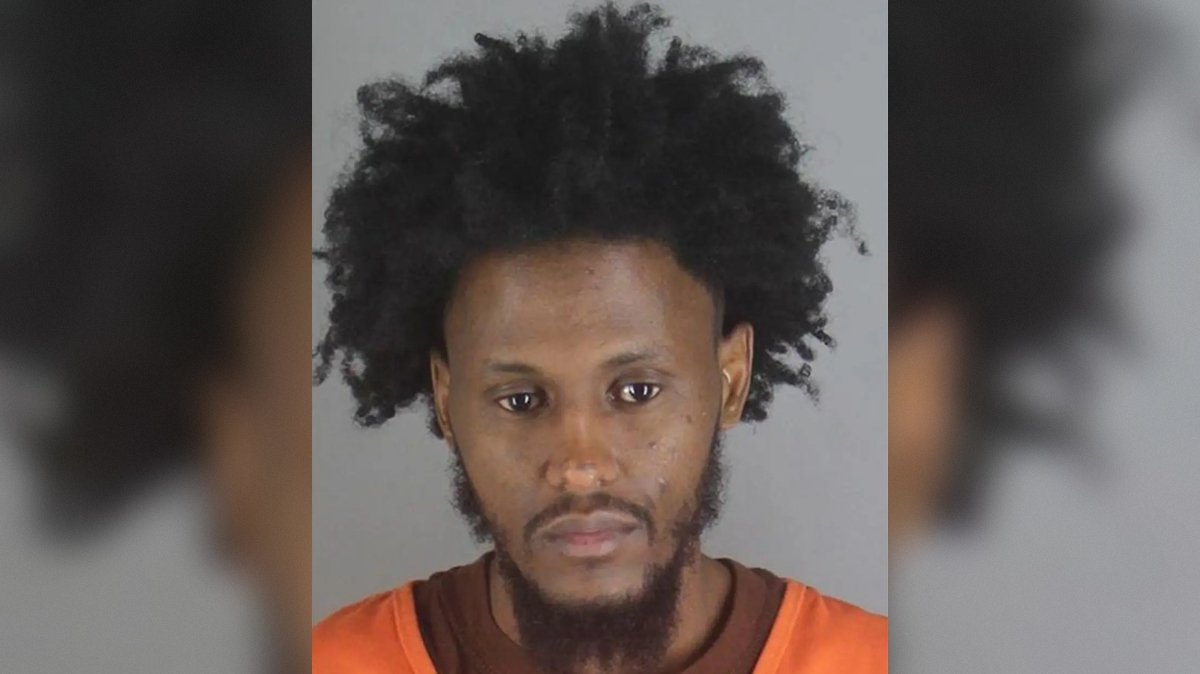Mugshot of a man with curly black hair wearing an orange prison uniform, looking directly at the camera.