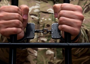 A handcuffed soldier behind the bars against a black background.