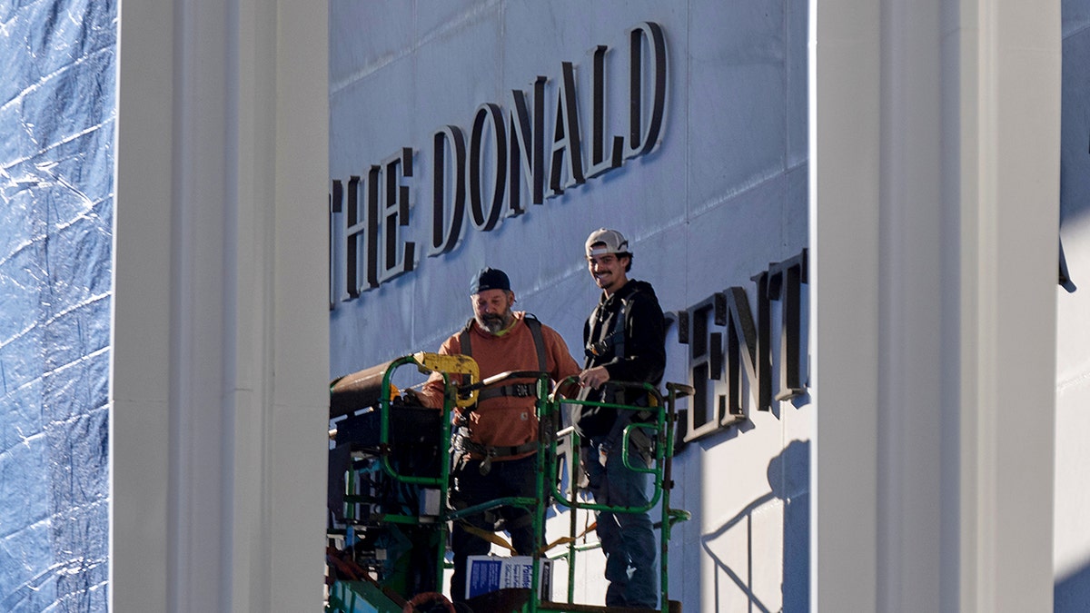 Two workers stand on a forklift beside 