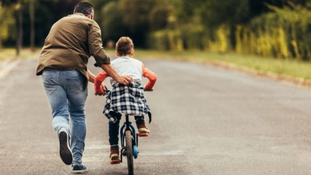 Father helping his child learn to ride a bicycle on a road.