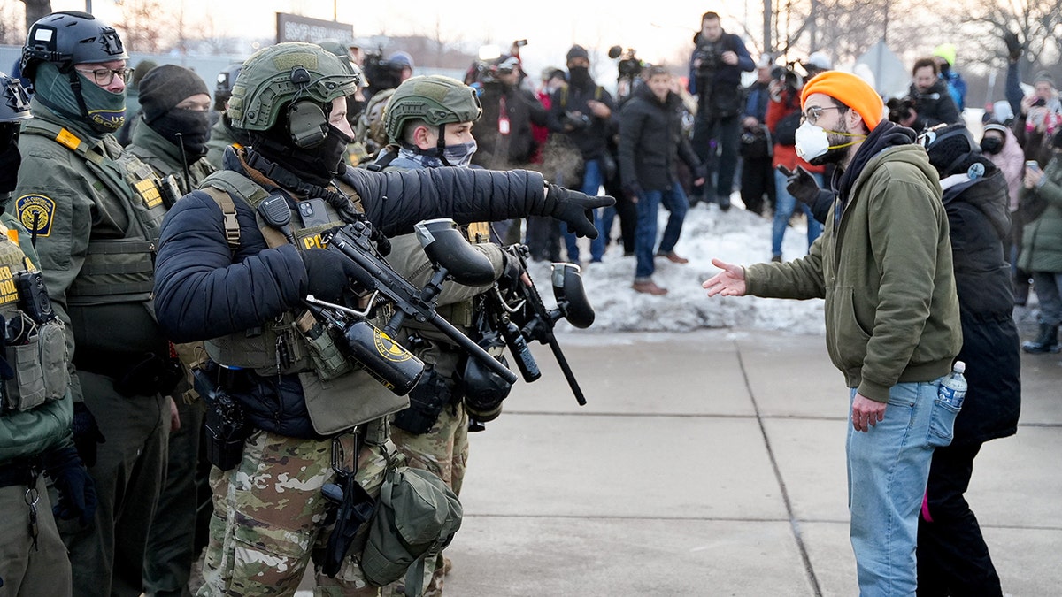 U.S. Border Patrol agents in tactical gear face a masked protester during a tense confrontation on a sidewalk.