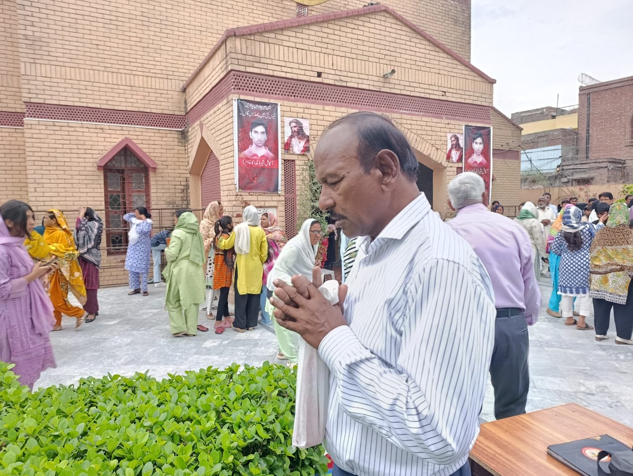 A man prays outside a church in Youhanabad, Lahore, Pakistan, on March 15, 2025, during commemorations marking the anniversary of the twin suicide bombings. Banners honoring Akash Bashir are visible in the background. | Credit: Kamran Chaudhry